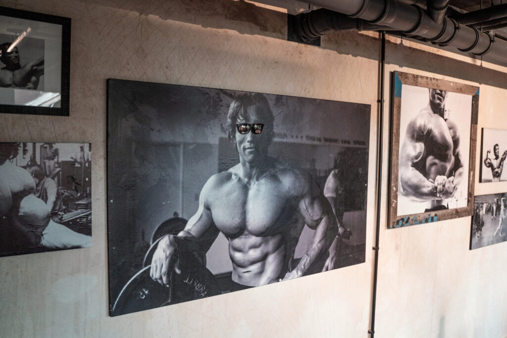 Bild von einem muskulösen Mann in Sonnenbrillen, der mit einer Gewichtscheibe posiert; um ihn herum hängen Schwarz-Weiß-Fotos, die Bodybuilding zeigen.