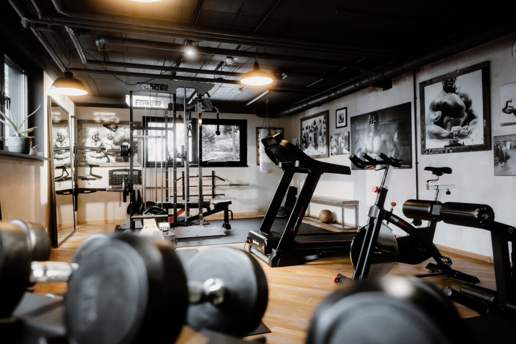 Fitnessraum mit verschiedenen Geräten, einschließlich einer Kraftstation und einem Laufband, umgeben von Sportfotografien.