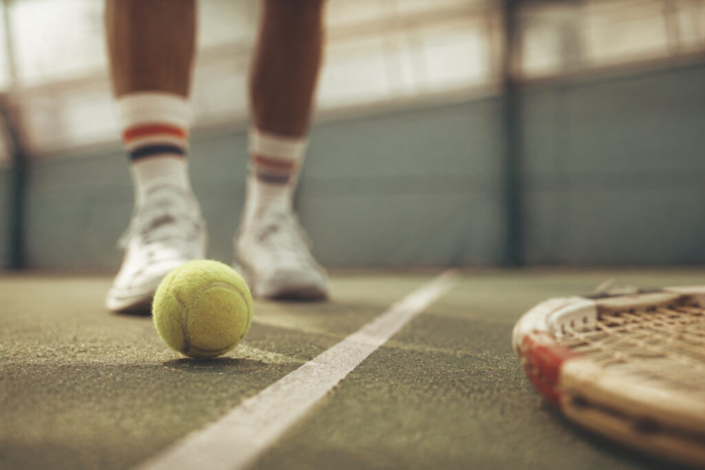 Bein eines Tennisspielers steht über einem Tennisball auf einem Platz, links liegt ein Tennisschläger. Natürliches Licht.