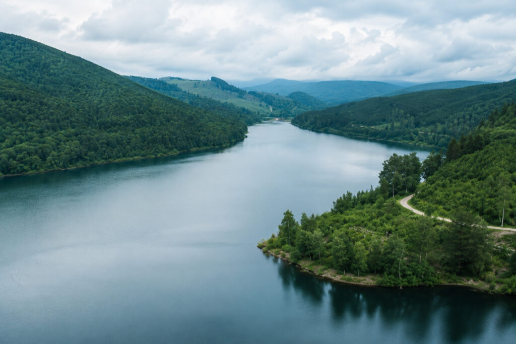 Blick auf einen ruhigen See, umgeben von hügeligen Wäldern und grünen Ufern unter einem bewölkten Himmel.