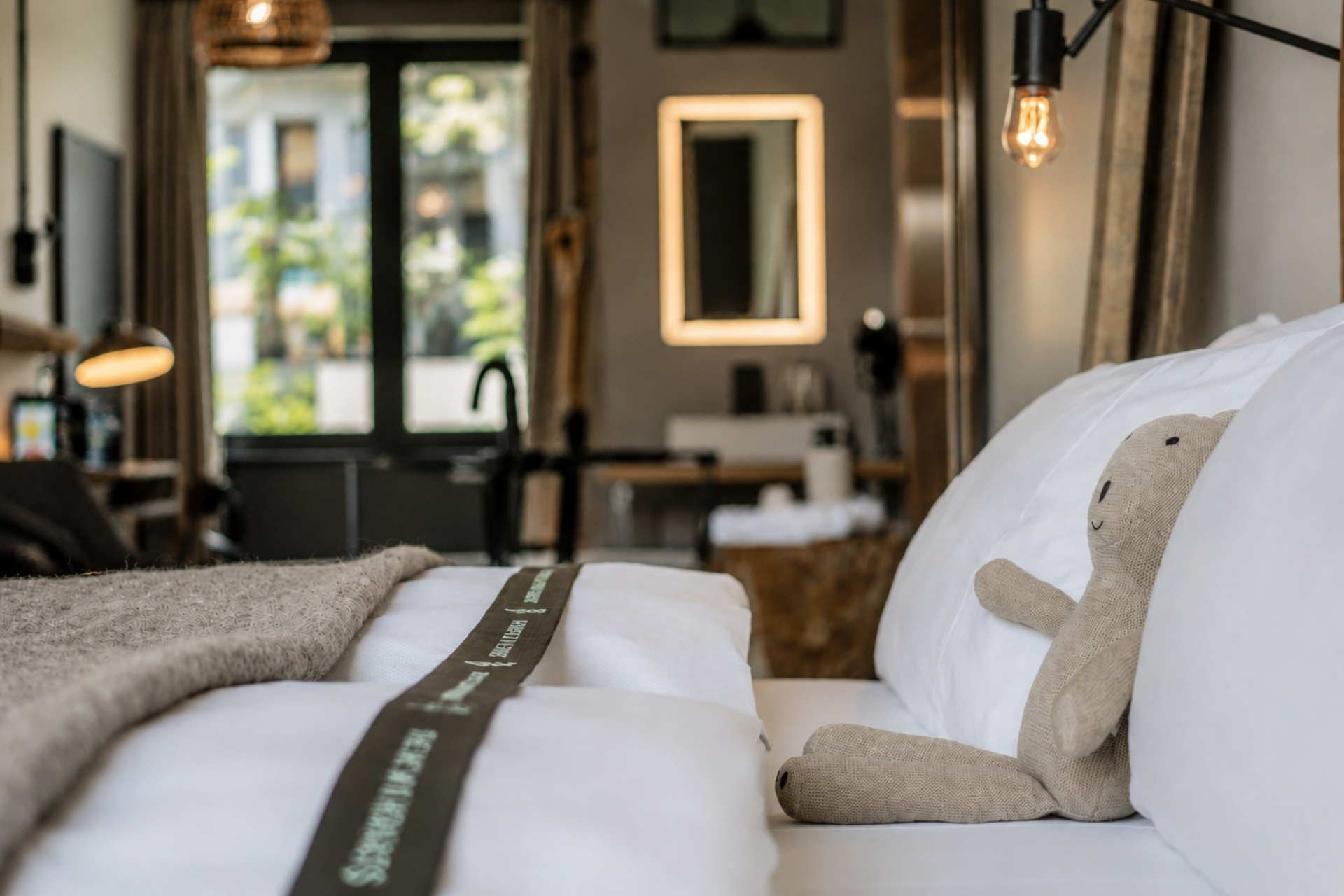 Gemütliches Schlafzimmer mit Bettdecke, zwei Kopfkissen und einem Stoffbären. Im Hintergrund Fenster und gemütliche Beleuchtung.