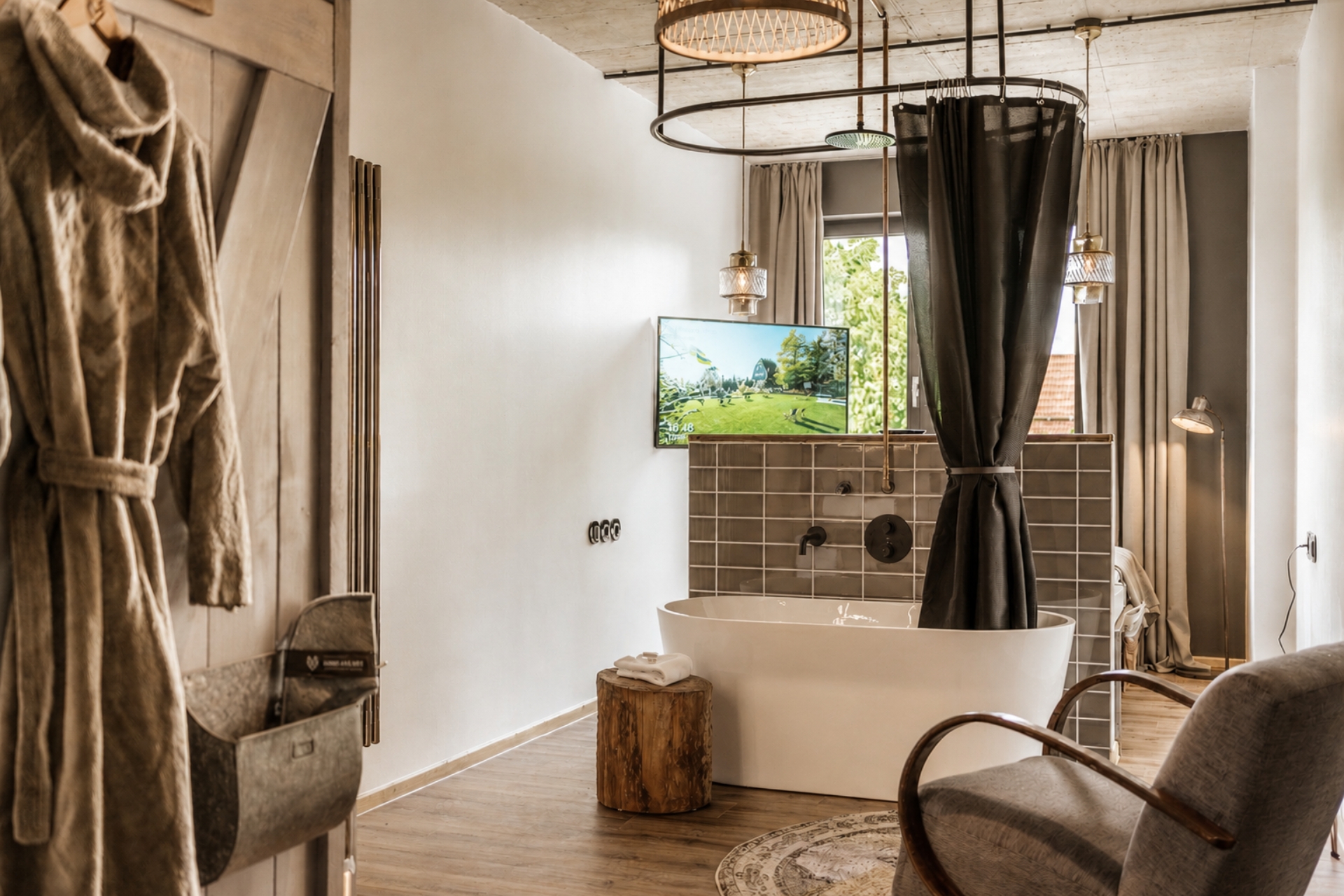 Modernes Badezimmer mit freistehender Badewanne, TV, Holzstuhl und textilem Badetuch. Helle Farben und große Fenster.