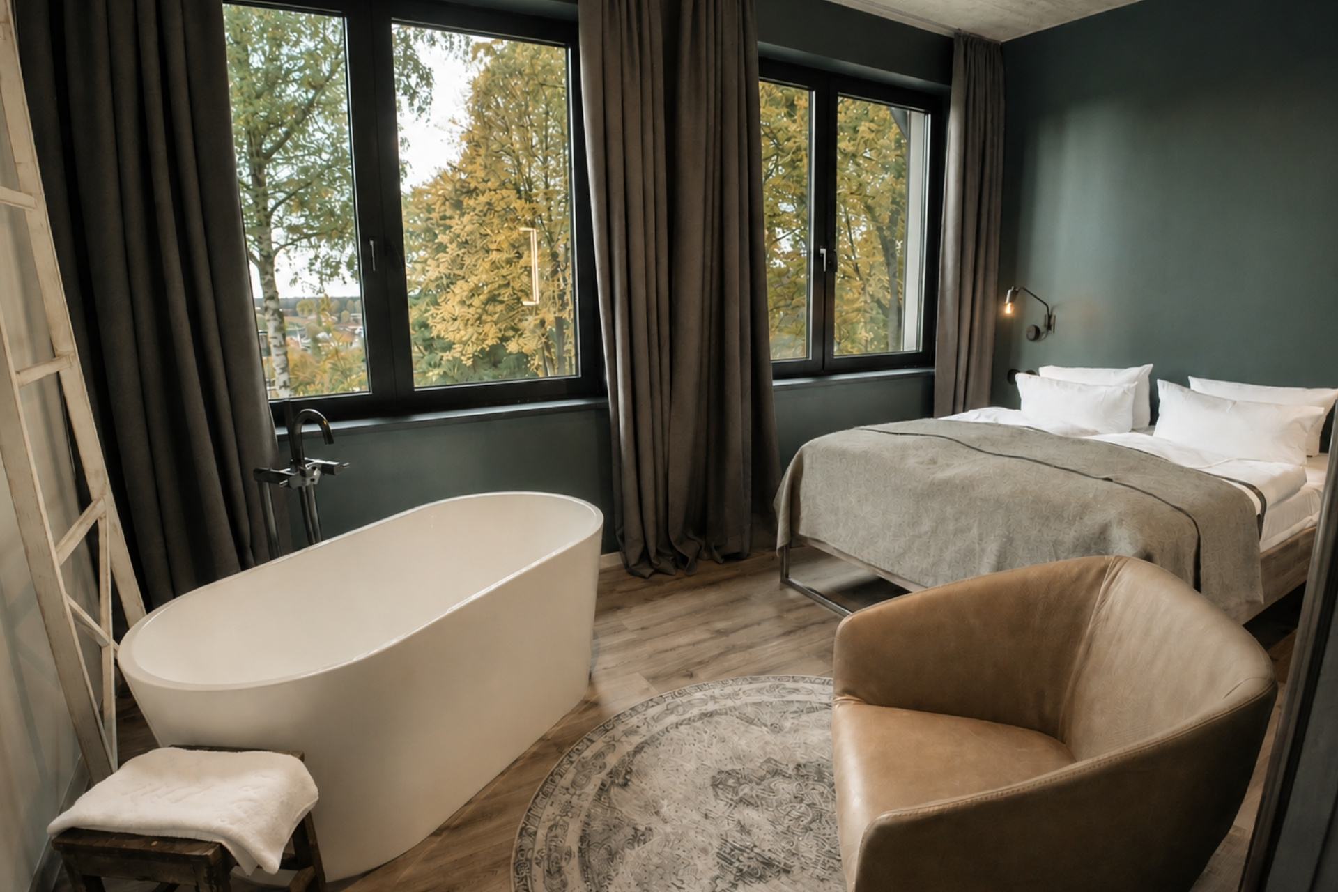 Helles Schlafzimmer mit doppeltbett, freistehender Badewanne, Sessel, Teppich und großen Fenstern mit Blick auf Bäume.