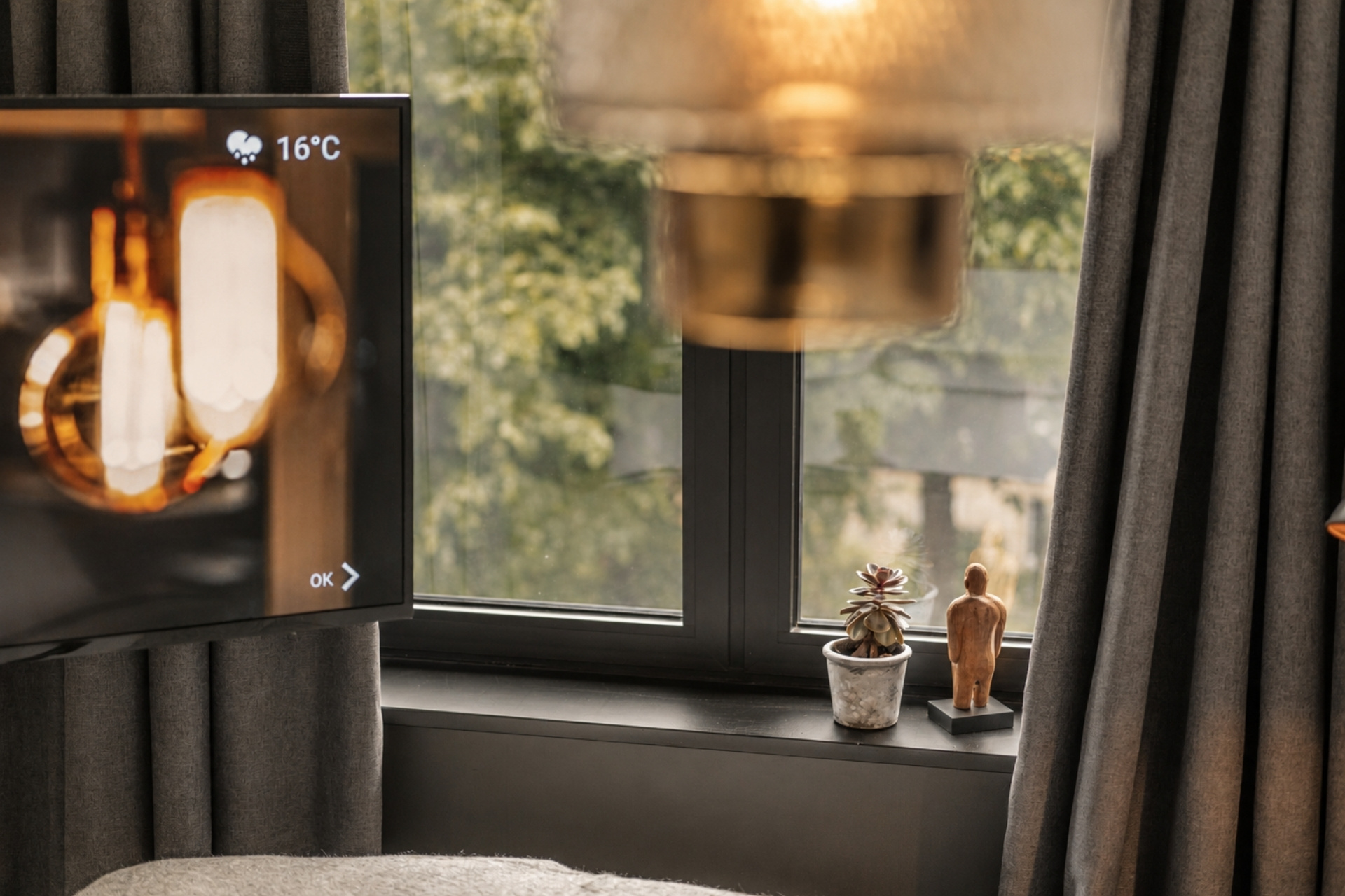 Wohnzimmer mit Fernseher, Temperaturanzeige, Pflanzen und Holzfigur auf der Fensterbank; Tageslicht und Vorhänge sichtbar.