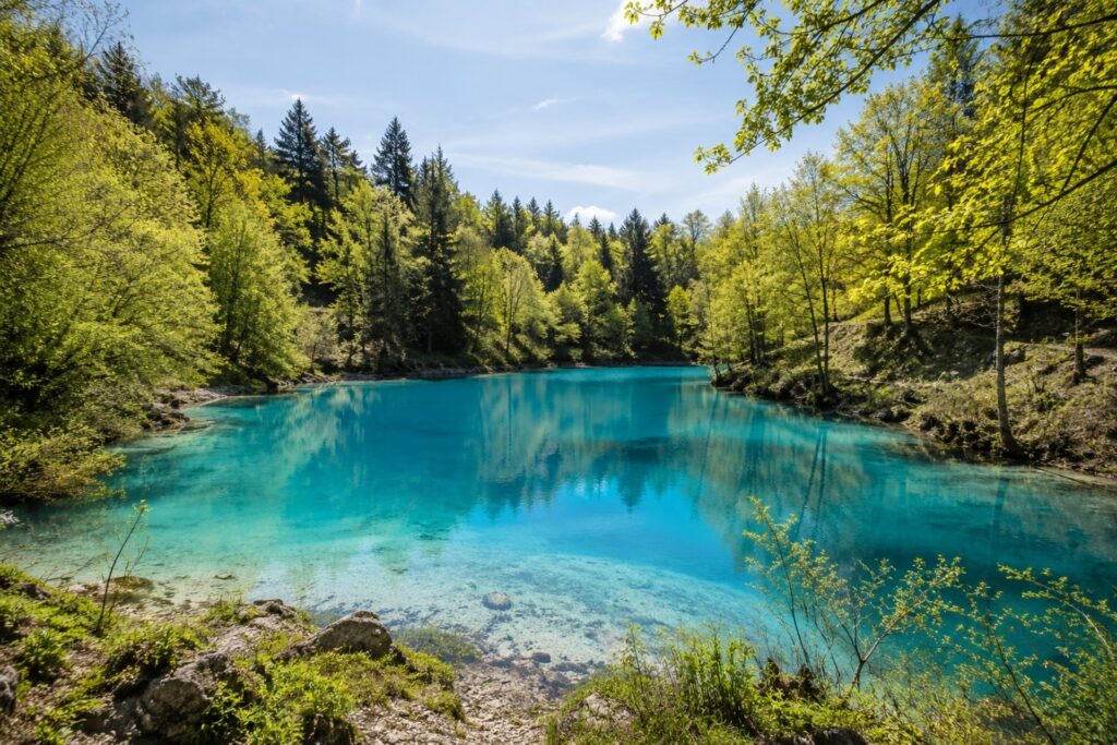 Türkisfarbener See umgeben von grünen Bäumen und Felsen unter blauem Himmel, reflektiert die natürliche Umgebung.