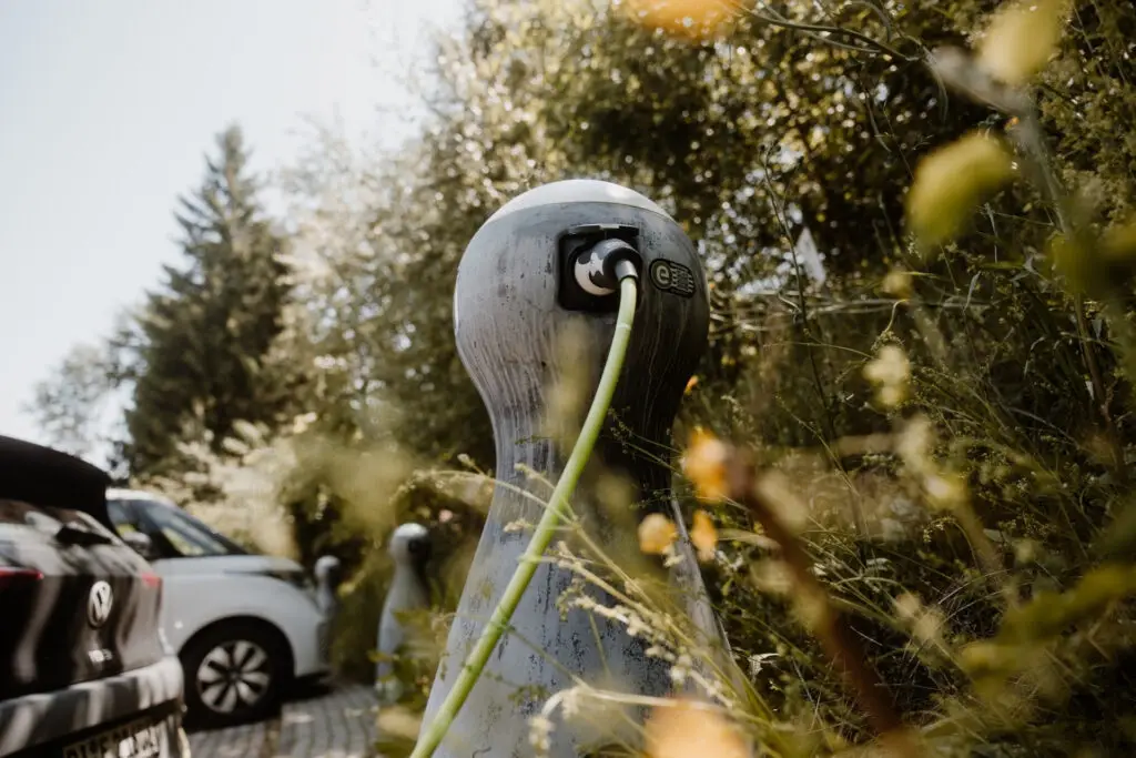 Ladestation für Elektrofahrzeuge im Freien, umgeben von Pflanzen und zwei parkenden Autos.