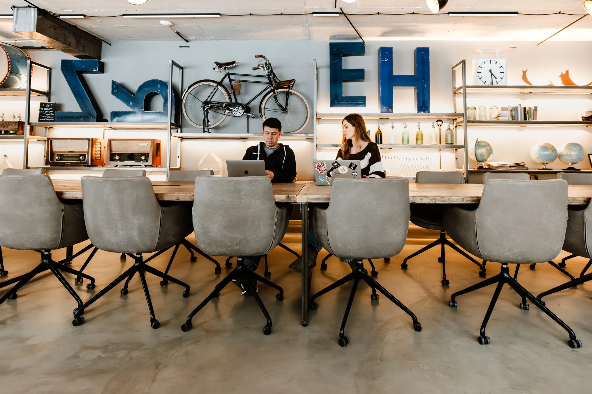 Zwei Personen arbeiten an einem Tisch in einem modernen Raum mit Regalen, Fahrrädern und Dekorationsobjekten im Hintergrund.