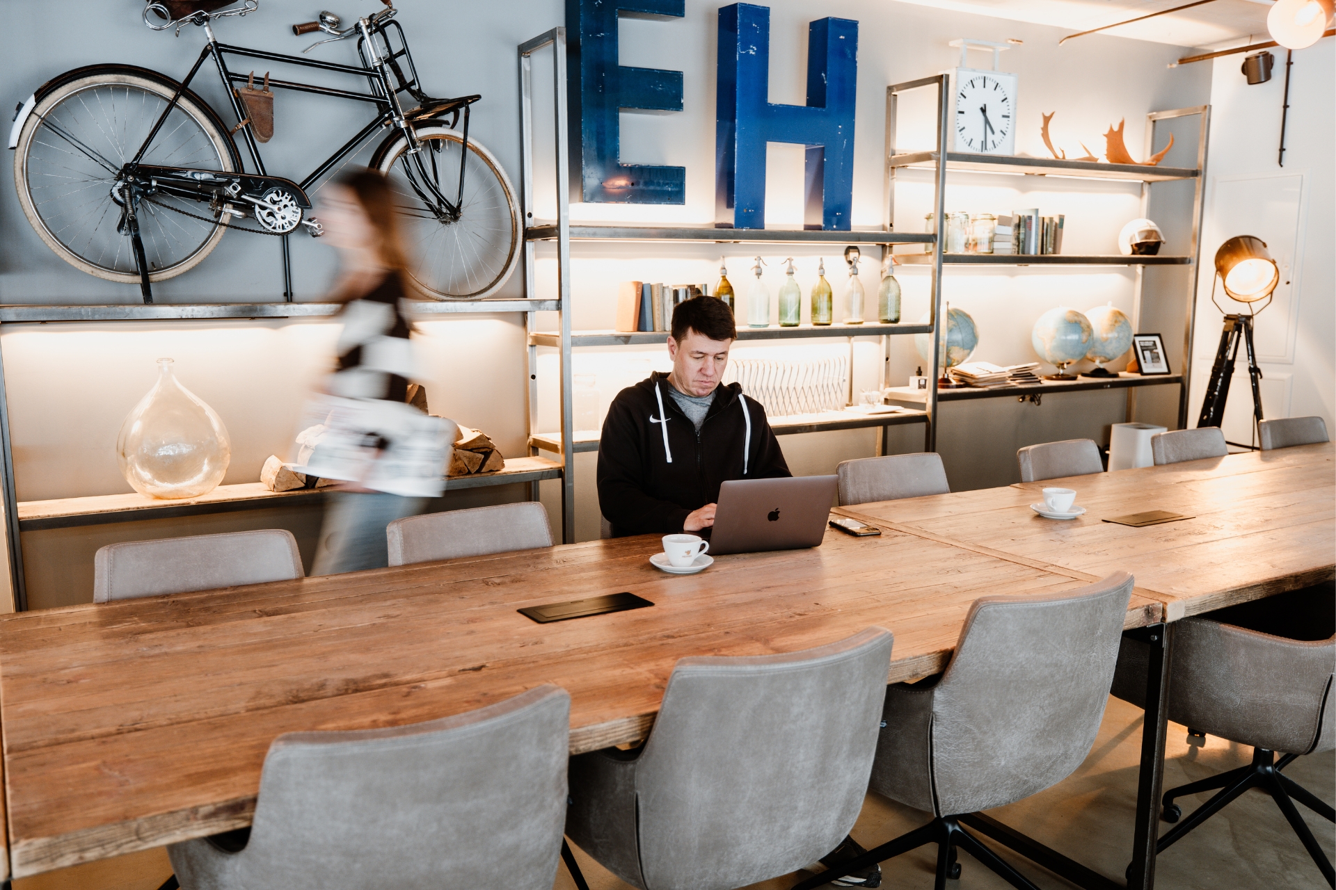 Zwei Personen sitzen an einem langen Tisch in einem modernen Raum, arbeiten an Laptops, im Hintergrund Holzstapel und Dekoration.