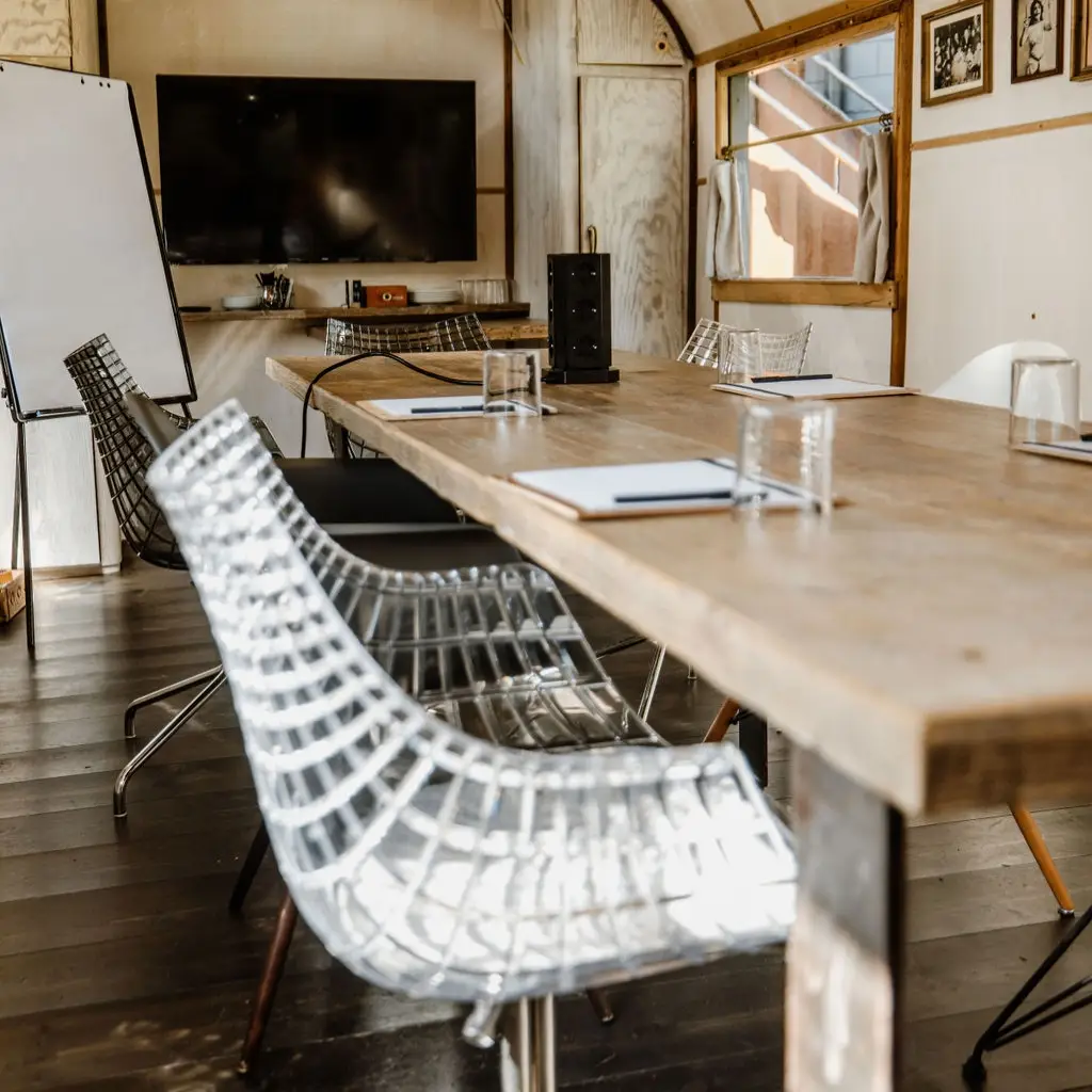 Besprechungsraum mit einem langen Holztisch, transparenten Stühlen, Monitor, Whiteboard und Notizmaterialien.