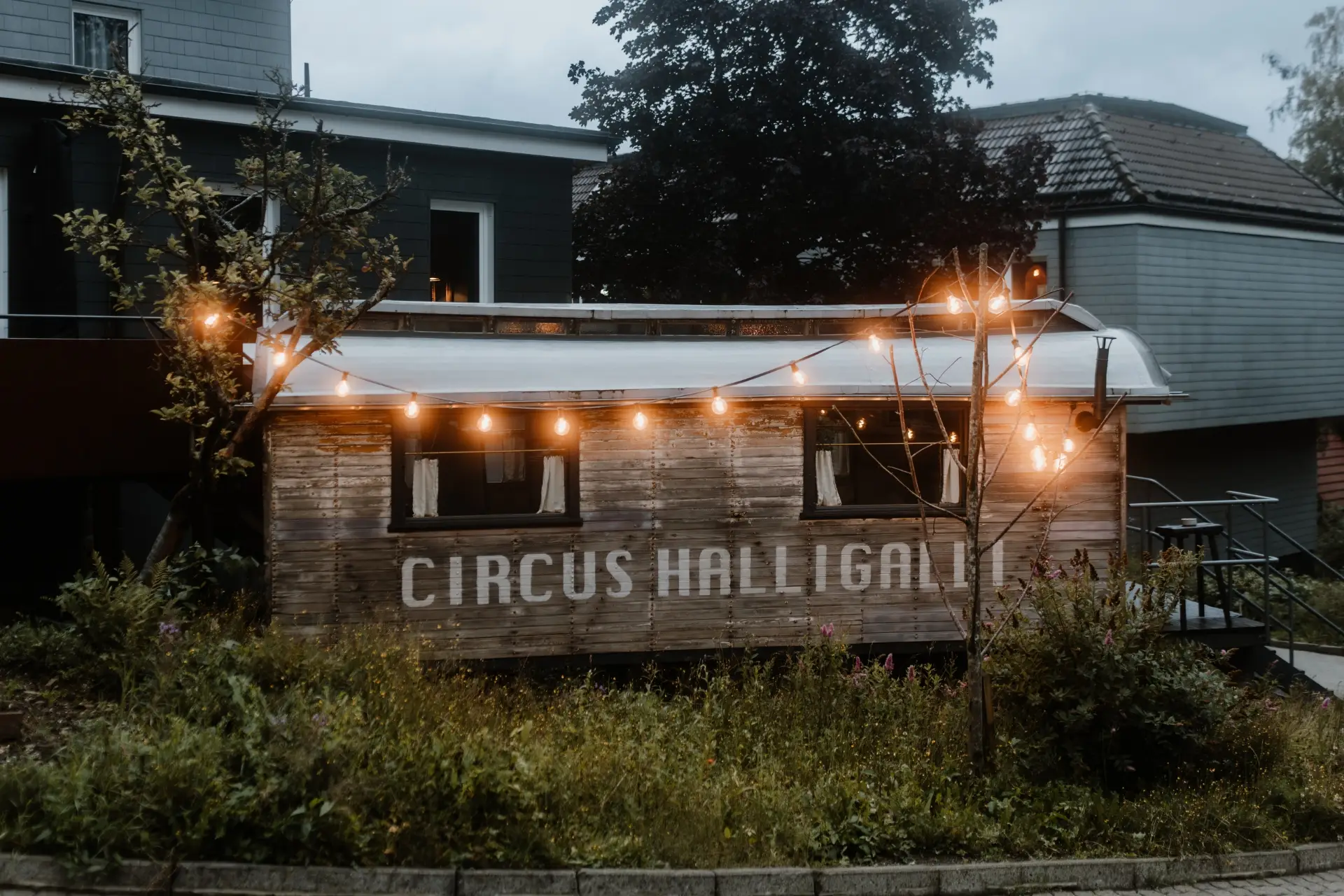 Holzhütte mit der Aufschrift „Circus Halligalli“, umgeben von Pflanzen und Lichtern, in einer städtischen Umgebung bei Dämmerung.