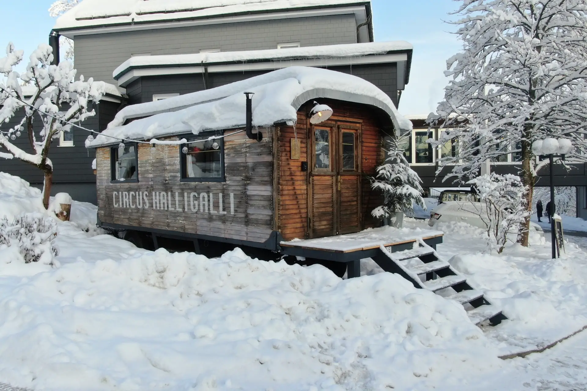 Holzhütte mit der Aufschrift CIRCUS HALiGALLi umgeben von Schneedecke, neben einem modernen Gebäude und schneebedeckten Bäumen.