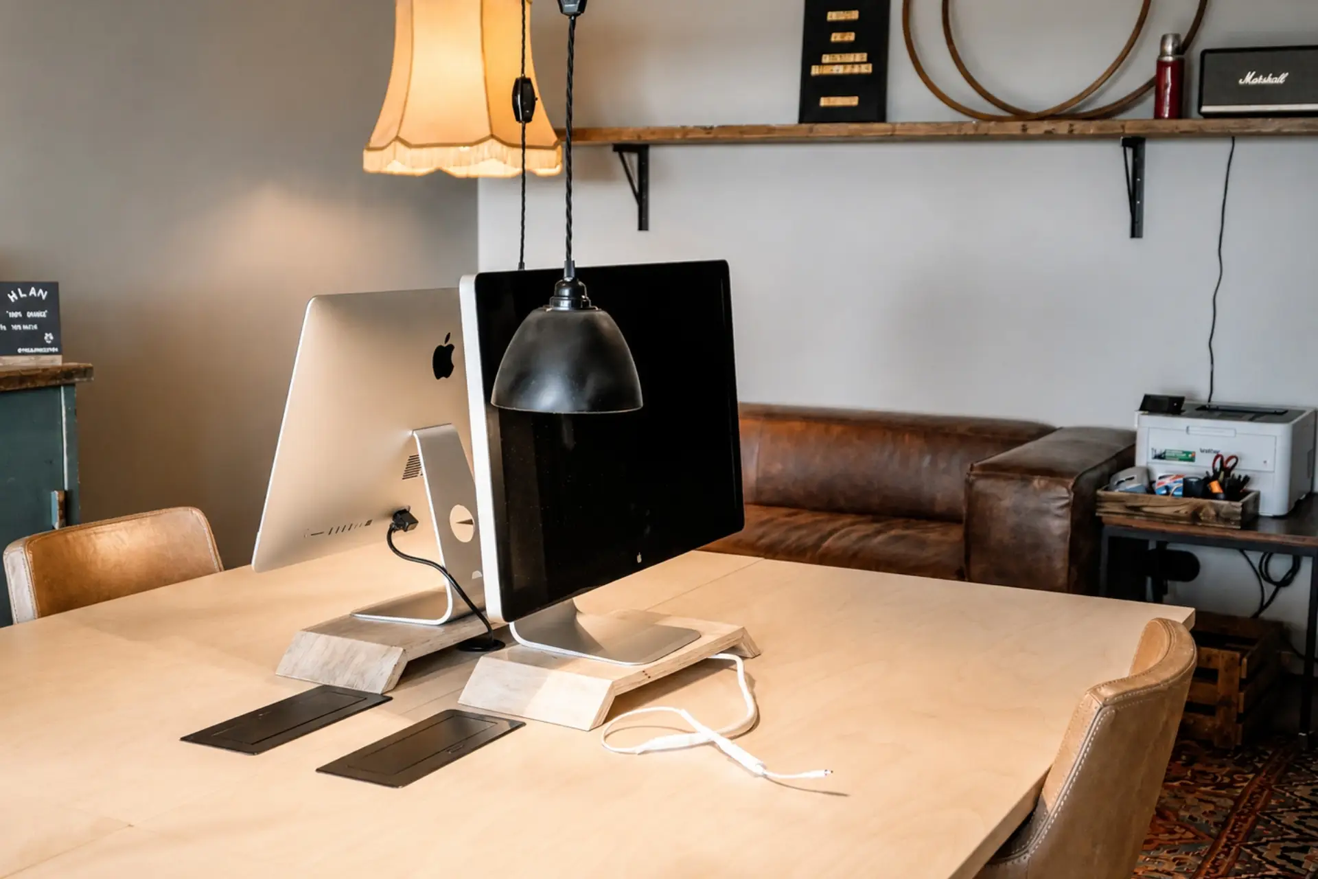 Schreibtisch mit zwei iMacs, einer Pendelleuchte und einem Ledersofa im Hintergrund, geeignet für kreatives Arbeiten.