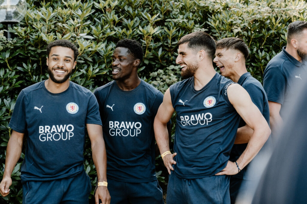 Fünf Lächelnde Männer in Sportbekleidung mit BRAVO GROUP-Aufschrift stehen zusammen vor einer Hecke.