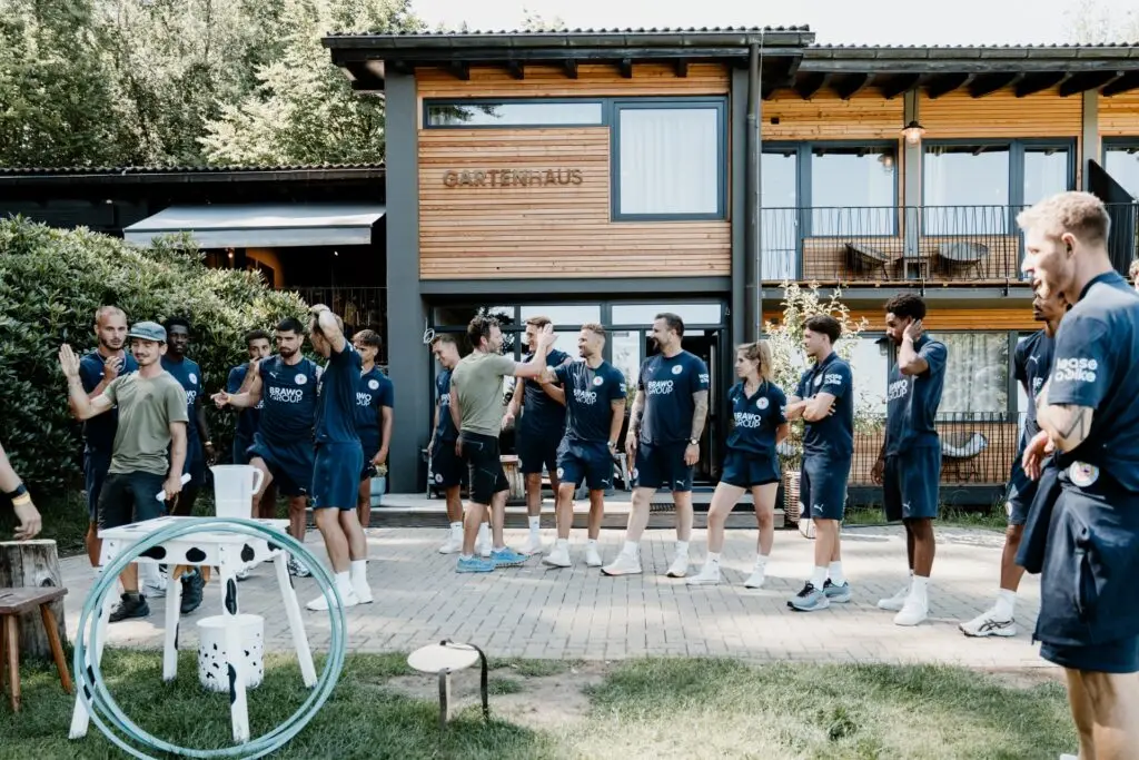 Gruppe von Sportlern in blauen Trikots steht vor einem Holzgebäude, bereit für eine Besprechung im Freien.