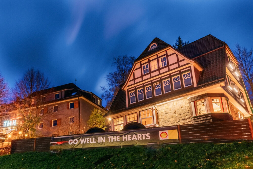 Historisches Fachwerkhaus bei Dämmerung mit Banner „GO WELL IN THE HEARTS“ im Vordergrund. Umgebende Bäume und Beleuchtung sichtbar.