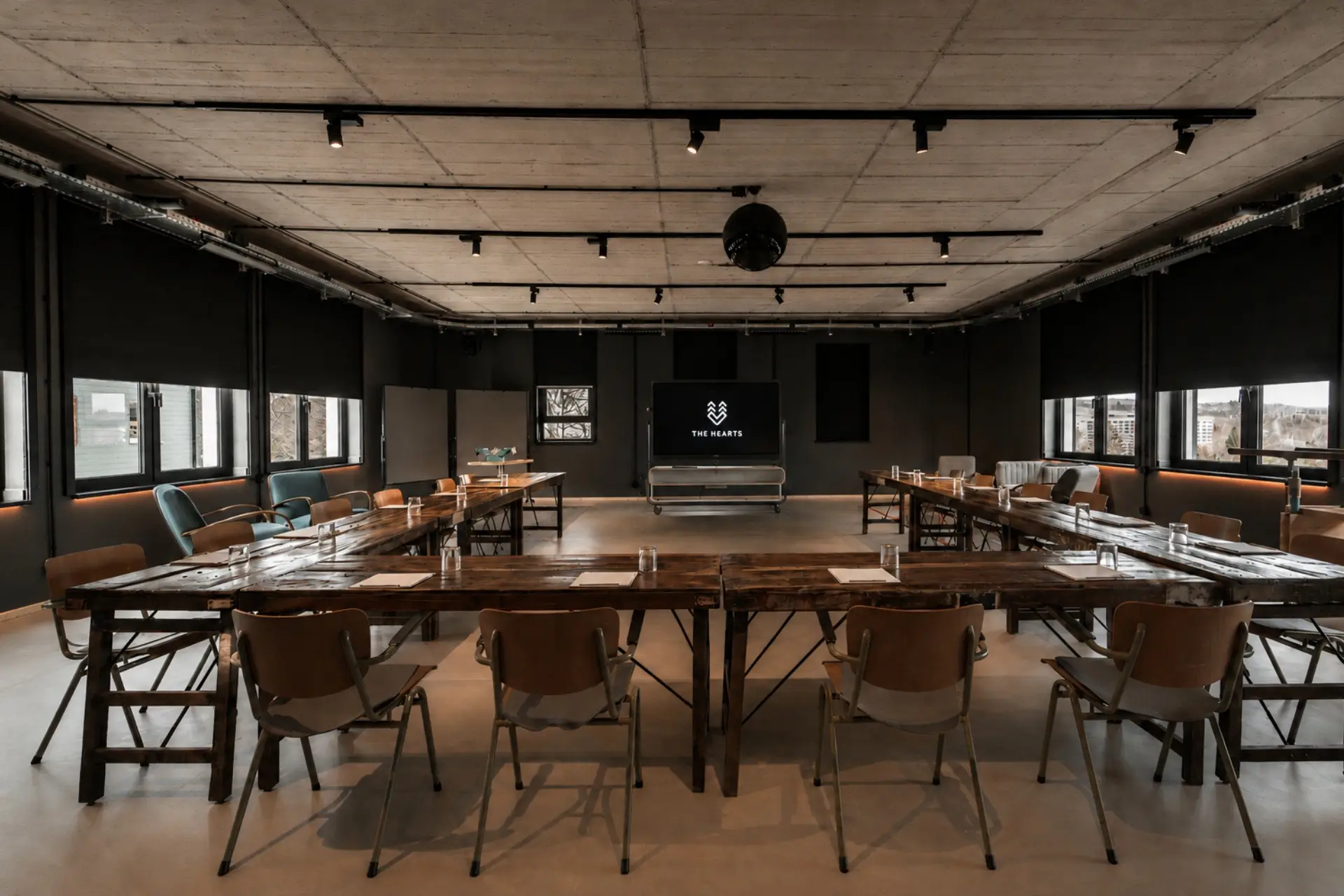 Konferenzraum mit langen Holztischen, Stühlen und einem Bildschirm im Hintergrund; moderne, minimalistische Einrichtung.