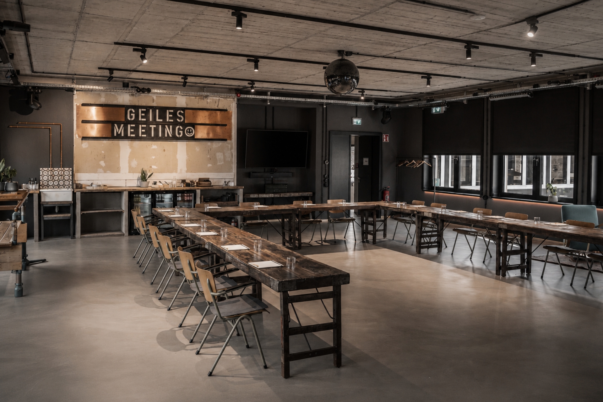 Heller Meetingraum mit Holztischen, Stühlen und einer Wand mit der Aufschrift GEILES MEETING. Fernseher an einer Wand.