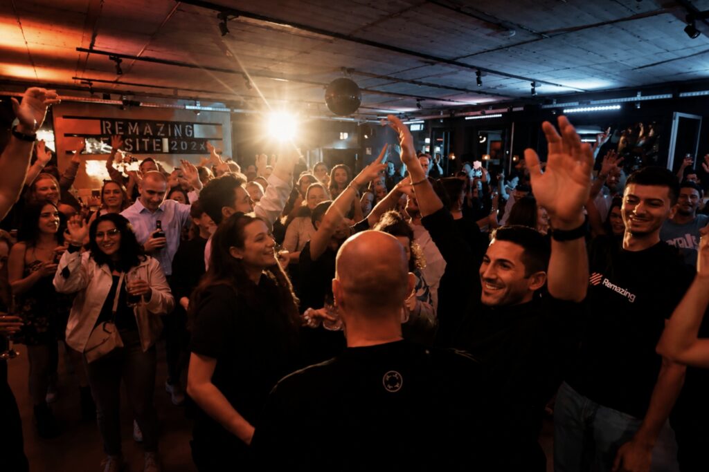 Menschen feiern in einem lebhaften Raum, heben Hände und lächeln, umgeben von Musik und bunten Lichtern.