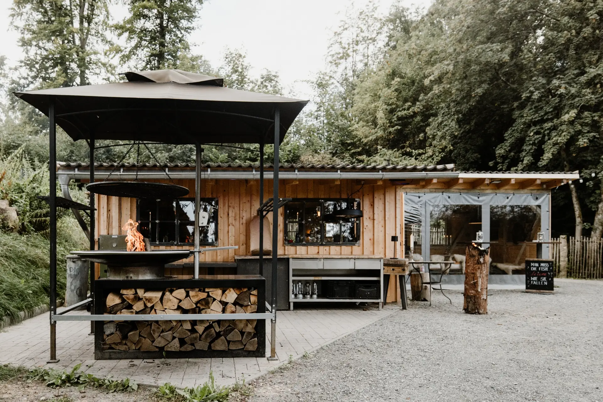 Hütte im Freien mit Holzscheiten, Grill mit offener Flamme und angrenzendem Holzgebäude, umgeben von Bäumen.