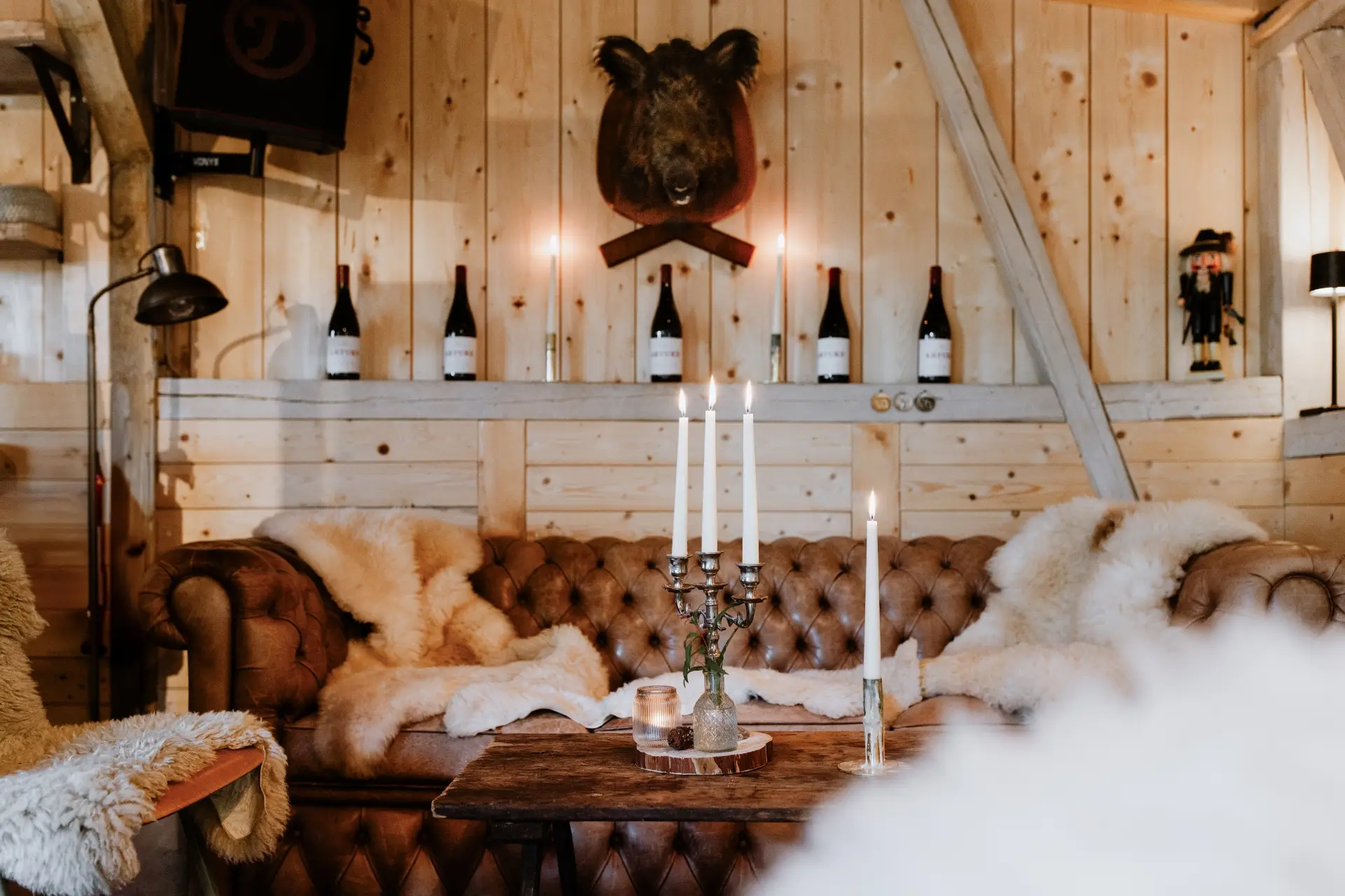 Gemütliche Ecke mit braunem Ledersofa, Holztisch, Kerzen und Weinflaschen an der Wand in holzverkleideter Umgebung.