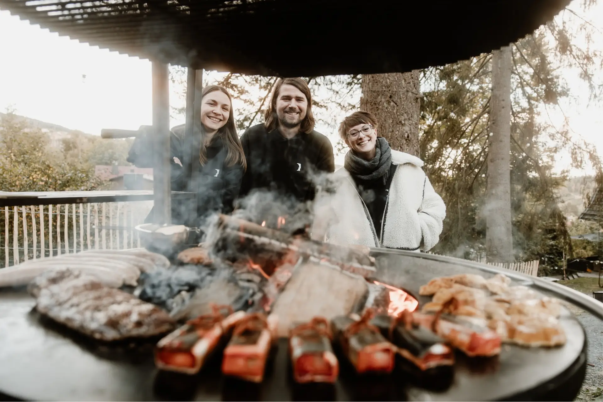 Drei Personen stehen lächelnd vor einem Grill mit Fleisch und Gemüse in einer natürlichen Umgebung.