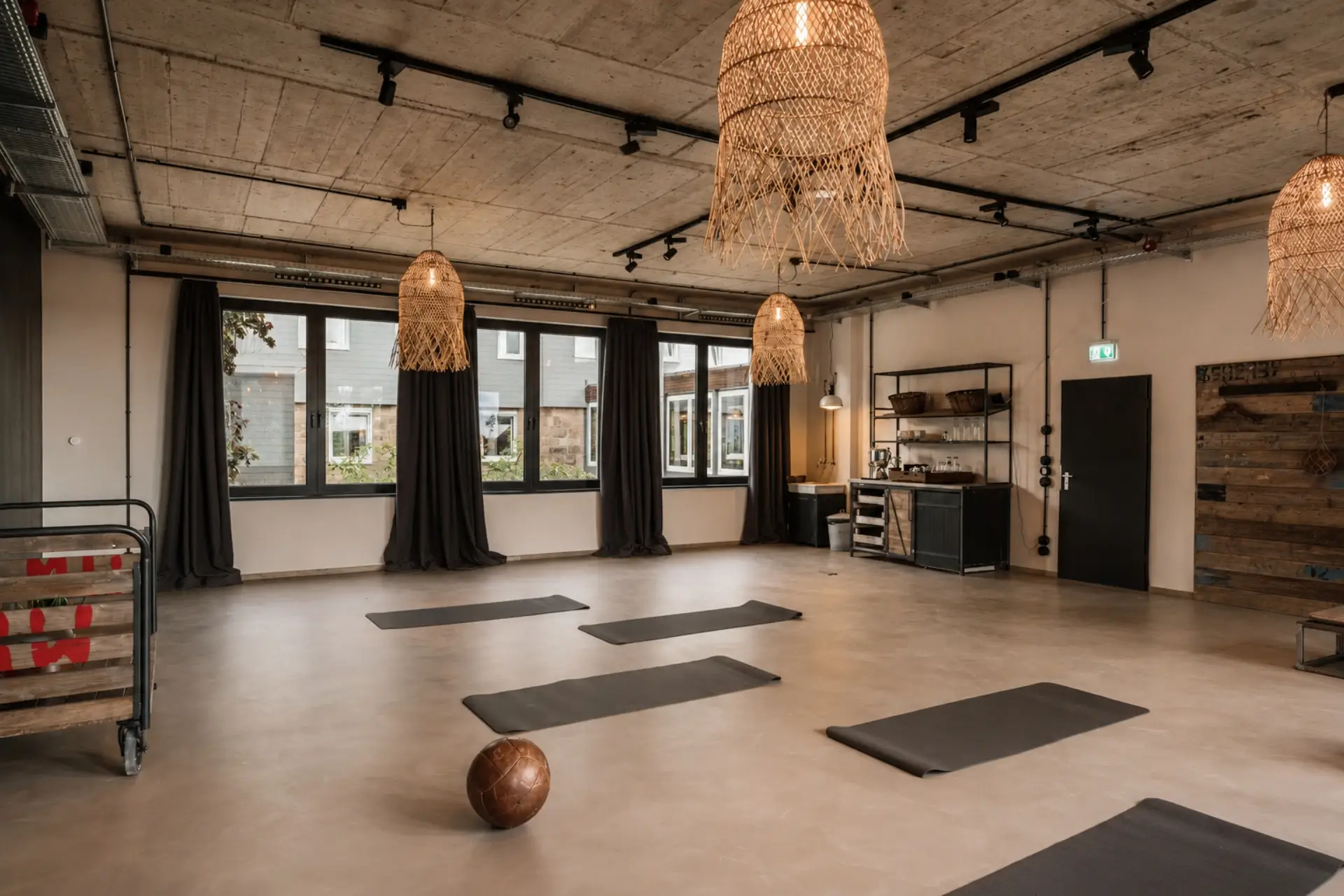 Heller Raum mit Holzdekor, Yogamatten am Boden, großen Fenstern und geflochtenen Lampen an der Decke.
