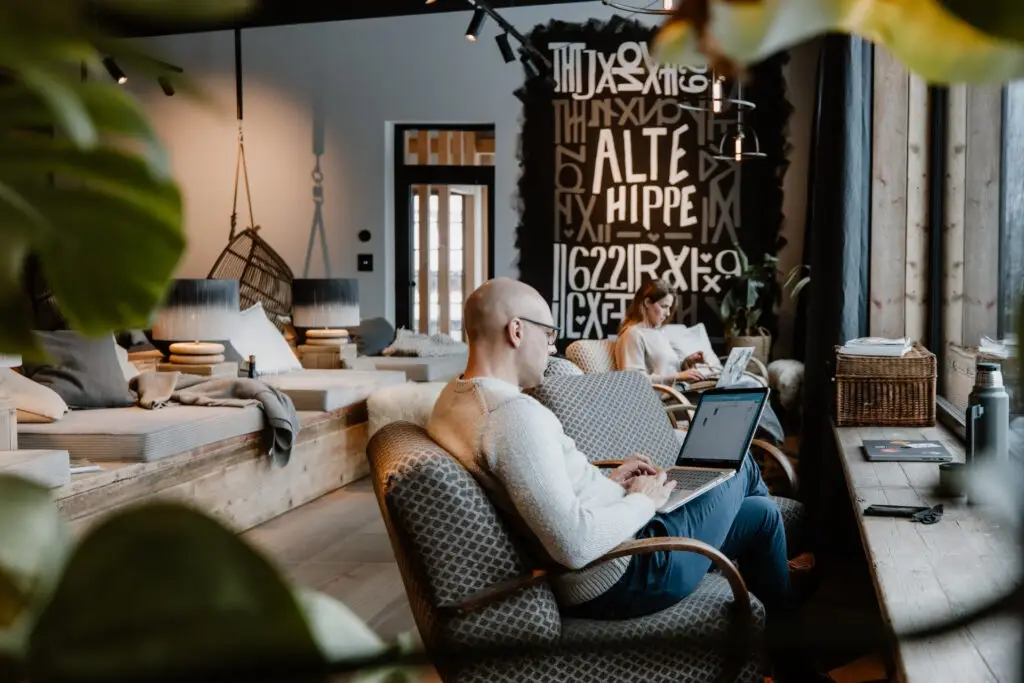 Ein Mann sitzt am Laptop und arbeitet in einem stilvollen, hellen Café. Eine Frau im Hintergrund nutzt ebenfalls einen Laptop.