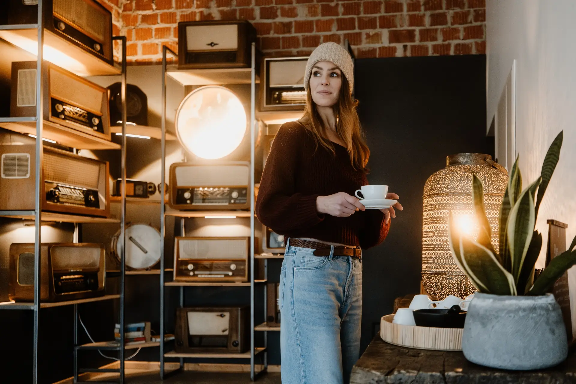 Frau mit Kaffeetasse steht in einem Raum mit Vintage-Radios, dekoriert mit Pflanzen und einer leuchtenden Lampe.