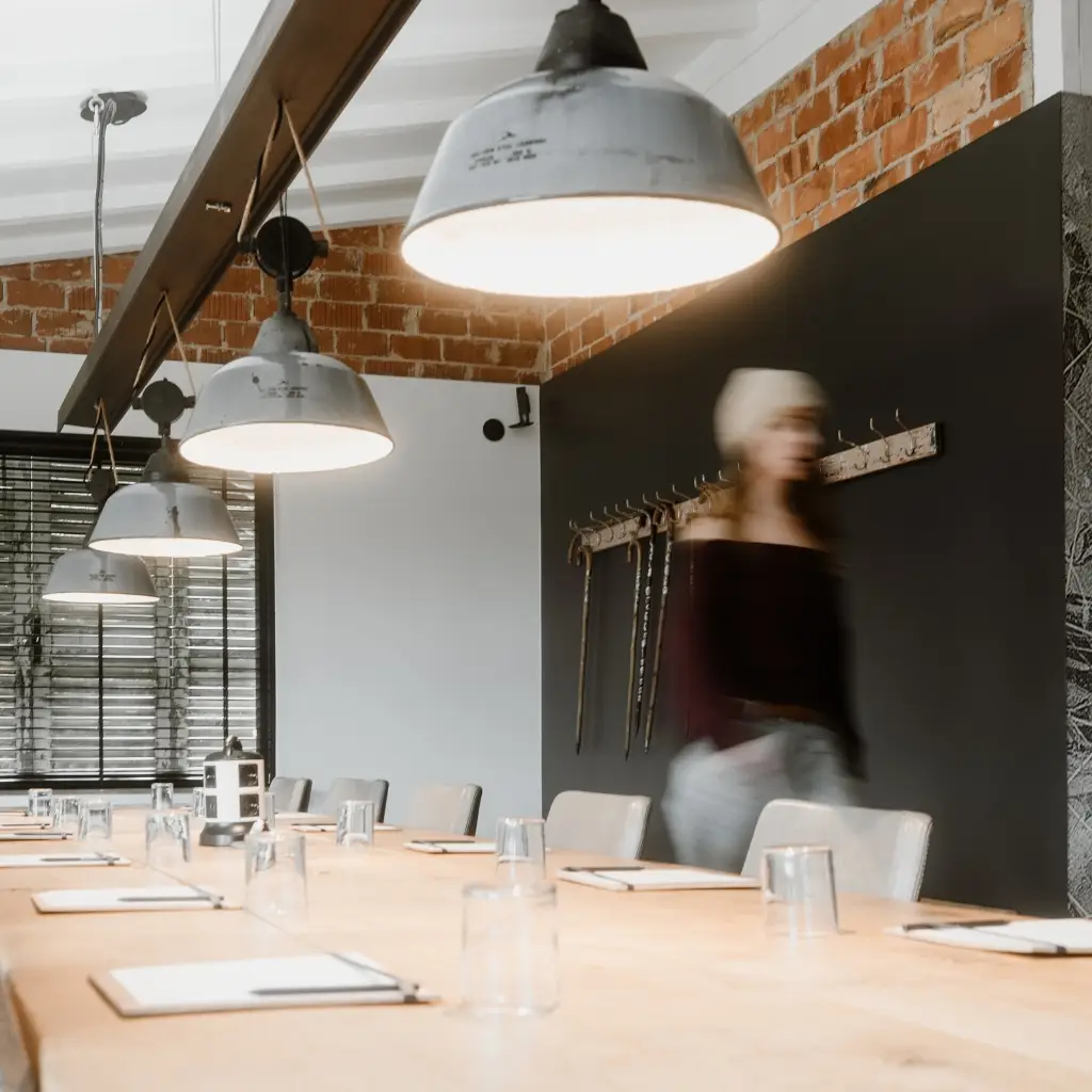 Frau mit Kopfbedeckung bewegt sich durch einen modernen Besprechungsraum mit Holz- und Betontisch und industrieller Beleuchtung.