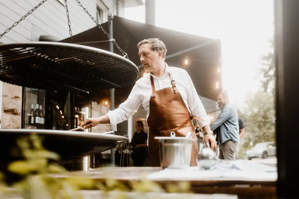 Ein Mann mit Schürze grillt im Freien, während im Hintergrund weitere Personen und eine Restaurantatmosphäre sichtbar sind.