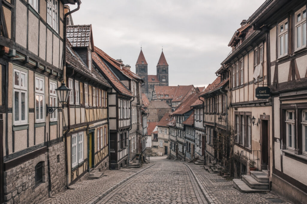Verlassene Kopfsteinpflasterstraße mit Fachwerkhäusern und einem entfernten Kirchturm im Hintergrund. Bewölkter Himmel.