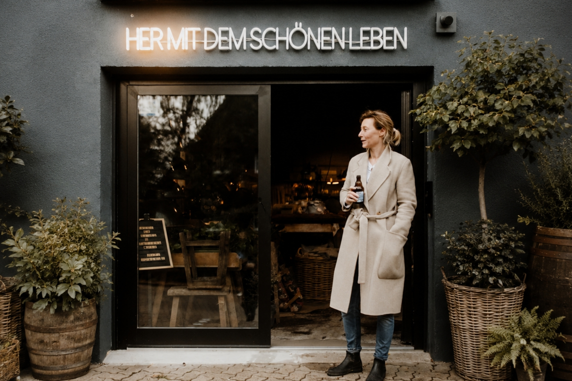 Frau in einem beigen Mantel steht vor einem dunklen Eingang mit einer Getränke-Label in der Hand, Pflanzen umgeben den Bereich.