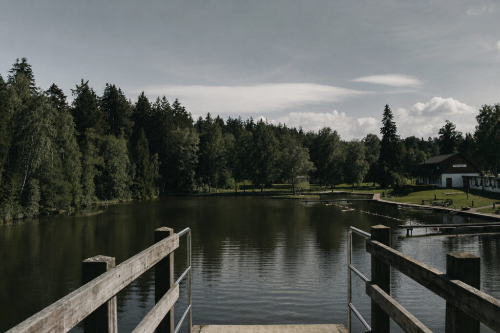 Fotografie eines ruhigen Sees mit Holzsteg, umgeben von Bäumen und einer Wiese. Im Hintergrund ist ein Gebäude sichtbar.
