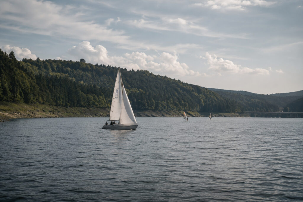 Segelboot mit mehreren Personen fährt auf ruhigem Wasser, umgeben von bewaldeten Hügeln und einer bewölkten Himmel.