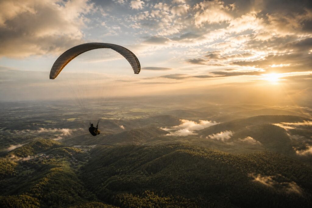 Paraglider schwebt über grüne Hügellandschaft bei Sonnenuntergang, mit sanften Wolken und einer klaren Aussicht.